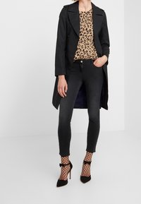 Žena oblečená v černém trenčkotě, topu s leopardím potiskem, černých slim kalhotách, rybářských ponožkách do kotníků a černých lodičkách s mašlemi.