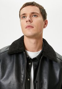 Chaqueta de piel negra con un suave cuello de borrego negro, que presenta una textura elegante, botones a presión y un cierre de cremallera, llevada sobre una camisa blanca.