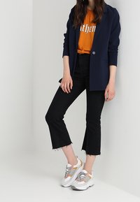 Femme portant un blazer bleu marine, un t-shirt graphique orange, un jean noir raccourci et des baskets blanches épaisses, debout contre un mur blanc.