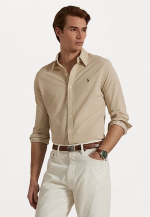 Beige long-sleeve button-up shirt with a subtle texture and embroidered logo. Paired with white pants and a brown belt, featuring a green watch.