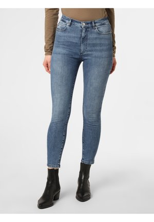 Vrouw draagt een high-waisted blauwe skinny jeans, een bruin shirt met lange mouwen en zwarte enkellaarsjes tegen een effen witte achtergrond.
