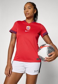 Maillot de football rouge avec des accents bleus et des manches à motifs, arborant un logo. Assorti à un short blanc et un ballon de football à motifs noir et blanc.