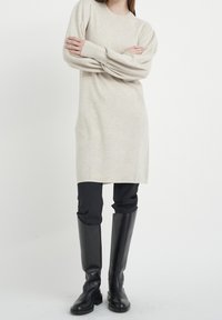 Robe pull beige tricotée avec manches longues, encolure arrondie et poignets côtelés, associée à un pantalon noir ajusté et des bottes noires montantes jusqu'aux genoux.