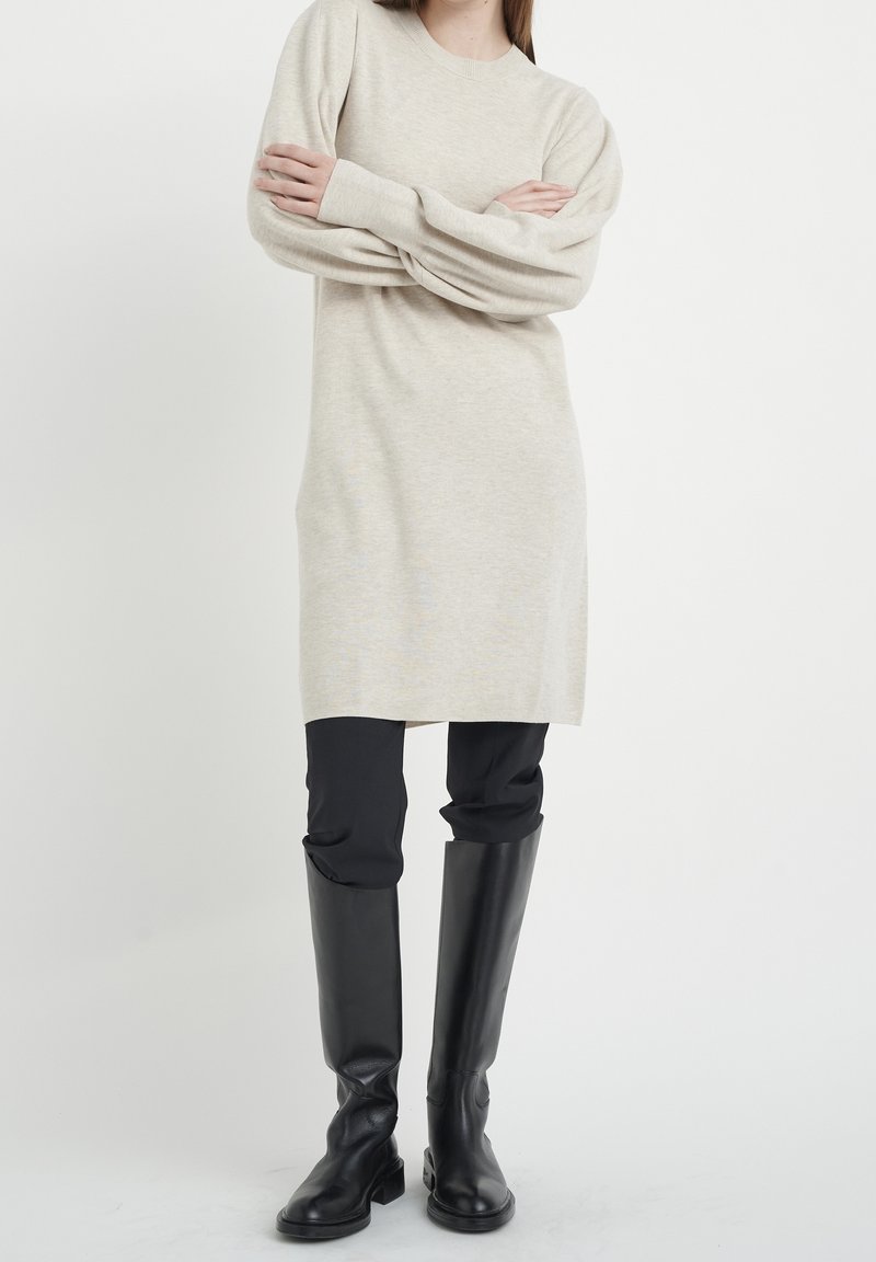 Robe pull beige tricotée avec manches longues, encolure arrondie et poignets côtelés, associée à un pantalon noir ajusté et des bottes noires montantes jusqu'aux genoux.