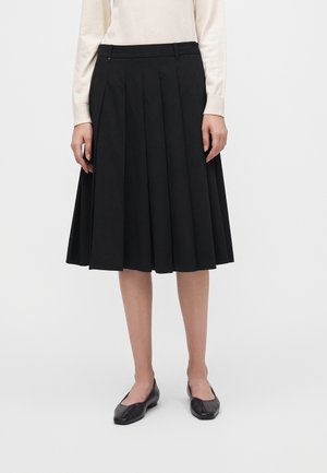 Vrouw draagt een zwarte geplooide knielange rok, een crèmekleurige top met lange mouwen en zwarte platte schoenen, staand tegen een effen achtergrond.