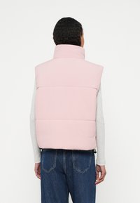 Růžová péřová vesta s vysokým límcem, prošitými panely a hladkou texturou, nošená přes světle šedé tričko s dlouhým rukávem a modré džíny.