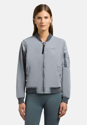 Kobieta w jasnoszarej bomberce z kieszeniami na zamek i ściągaczami na rękawach, w dopasowanych ciemnoszarych legginsach, stojąca na białym tle.