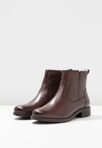 Une paire de bottes de cheville en cuir marron foncé avec une fermeture éclair latérale et un petit talon empilé sur une surface blanche et un fond gris clair.