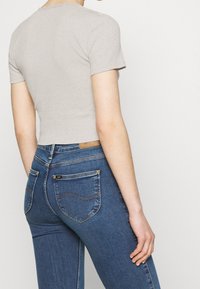 Šedý top s krátkým rukávem a vlnitým výstřihem, který je spárován s modrými skinny džíny s viditelným šitím a štítkem na zadní kapse.