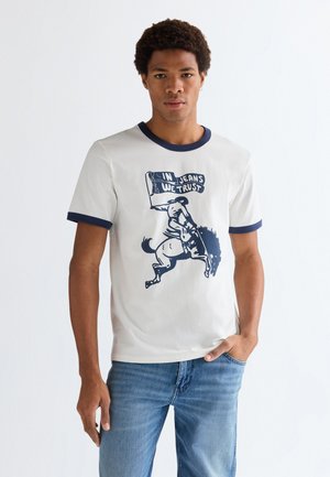 Mężczyzna ubrany w białą koszulkę z ciemnym obszyciem i grafiką jeźdźca na brykającym koniu trzymającego flagę z napisem "In Jeans We Trust", w połączeniu z niebieskimi dżinsami.