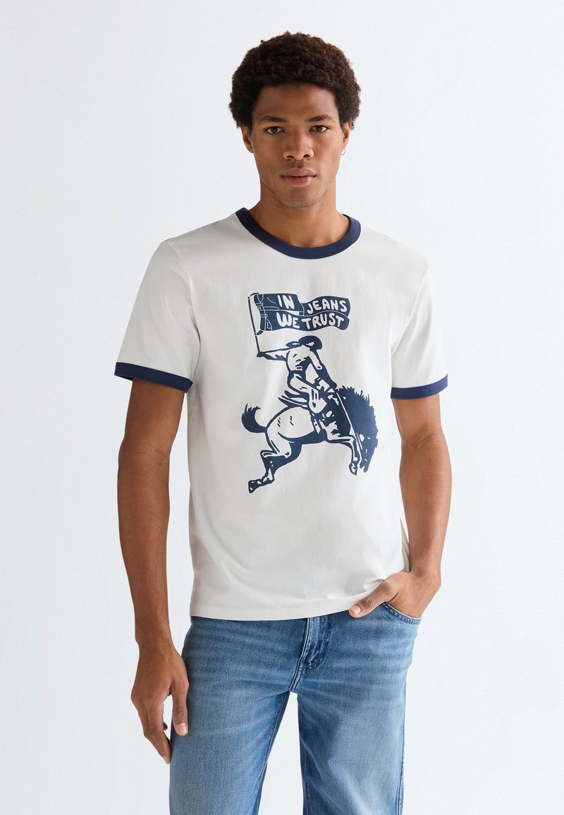 Άνδρας που φοράει λευκό μπλουζάκι με σκούρες λεπτομέρειες και εικόνα αναβάτη πάνω σε άλογο που σκαρφαλώνει, κρατώντας σημαία με την επιγραφή «In Jeans We Trust», σε συνδυασμό με μπλε τζιν.