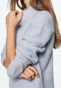 Maglione lavorato a maglia di colore grigio chiaro con una texture a costine, caratterizzato da un collo alto e polsini arrotolati. Materiale morbido e peloso, con una vestibilità comoda.