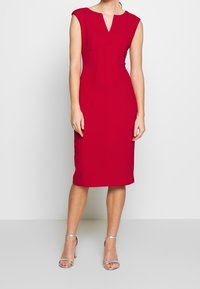 Robe sans manches rouge avec un décolleté en V et une silhouette ajustée, fabriquée en tissu lisse, atteignant le genou, accompagnée de sandales argentées à lanières.