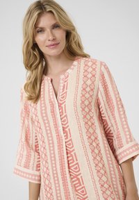 Tunic ușor în crem cu modele geometrice roz. Prezintă un guler tăiat și detalii cu nasturi, mâneci 3/4 și o croială lejeră.