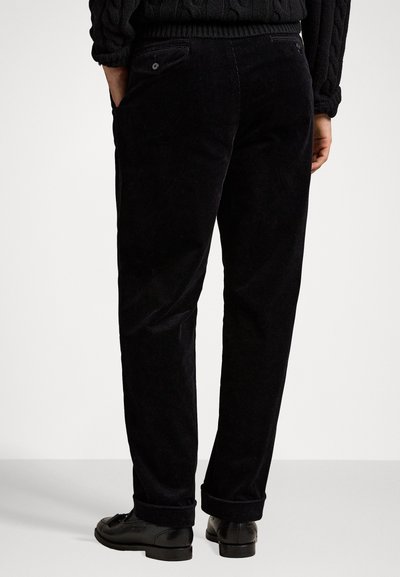 Pantalones de pana negros con una superficie texturizada, que presentan bolsillos traseros y dobladillos enrollados. Combinados con zapatos negros, mostrando un corte recto.