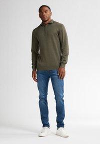 Olijfgroene sweater met rits, gebreide textuur, lange mouwen, gecombineerd met blauwe denim jeans en witte sneakers, staande tegen een neutrale achtergrond.
