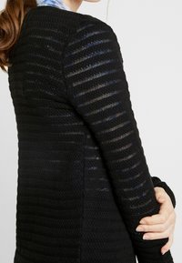 Černý pletený kardigan s texturovaným pruhovaným vzorem, dlouhými rukávy a fitted designem, předvádějící lehký a prodyšný materiál.