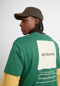 Grøn bomulds t-shirt med lysegule lange ærmer, har et hvidt rektangulært emblem med "Columbia" logoet på ryggen. Brun kasket.