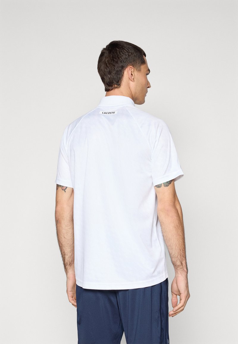 Polo blanc à manches courtes, avec un col et un logo Lacoste à l'arrière. Texture lisse, coupe standard, sans motifs visibles.