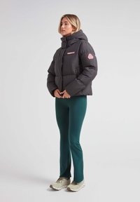Sort puffjakke med høj krave, pink branding og hexagonal patch. Båret over grønne leggings og beige sneakers. Glat tekstur.