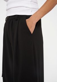 Schwarze hoch taillierte Hose mit glatter Textur, Seitentaschen, gefaltetem Vorderteil und einem dezent gestickten Logo in der Nähe des Bundes.