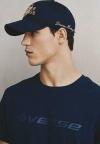 Granatowa czapka bejsbolowa z złotą haftowaną grafiką i czerwonymi akcentami; noszona z dopasowaną granatową koszulką z napisem "Diverse".