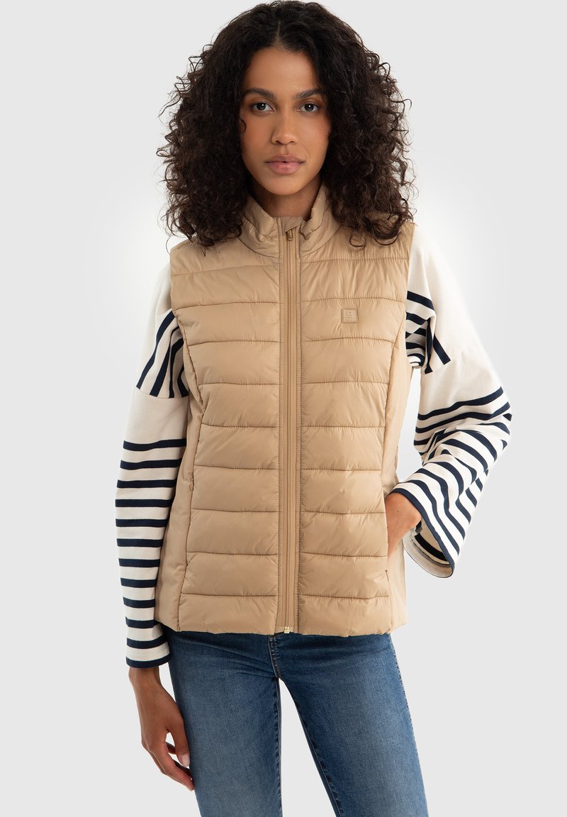 Bežinis pūkinis liemenė su vertikaliu siūlų raštu, aukštu apykakle ir priekiniais užtrauktukais. Dėvimas virš dryžuoto ilgomis rankovėmis marškinėlių, užbaigiant sluoksniuotą įvaizdį.