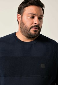 Navyblaues Strickpullover mit horizontaler Rippstruktur und einem schwarzen Logopatch auf der linken Brust. Rundhalsausschnitt mit einer lässigen Passform.