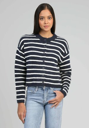 Jeune femme aux longs cheveux bruns portant un cardigan à boutons rayé bleu marine et blanc et un jean bleu clair, devant un fond gris uni.