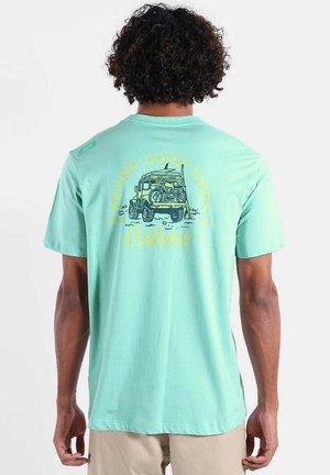 Personne portant un t-shirt vert clair avec un graphisme d'un véhicule vintage transportant un vélo, une planche de surf, et le texte « Having Good Times » au dos.