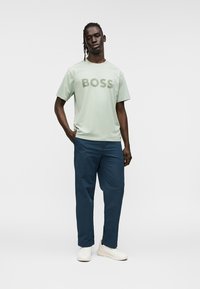Ανοιχτό πράσινο κοντομάνικο μπλουζάκι με την επιγραφή "BOSS" σε σκούρο πράσινο. Συνδυάζεται με σκούρο μπλε παντελόνι και λευκά αθλητικά παπούτσια. Λεία υφή υφάσματος, casual εφαρμογή.