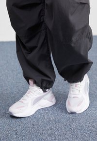 Zapatillas blancas con detalles en rosa y menta, que presentan paneles de malla, una suela acolchada y un acabado texturizado, combinadas con pantalones negros.