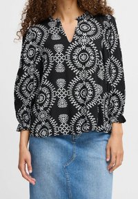 Blouse noire avec des motifs circulaires et floraux blancs, présentant un col en V et des poignets de manches froncés. Associée à une jupe en denim bleu.