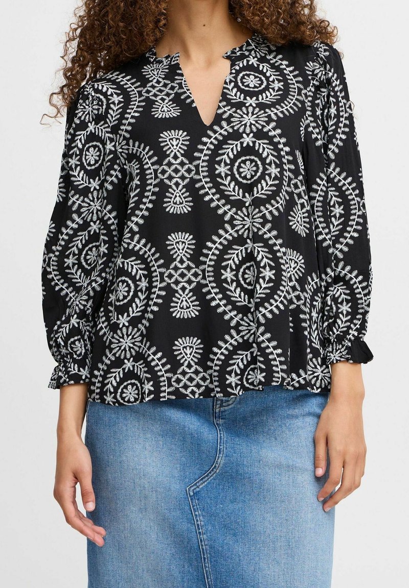 Blouse noire avec des motifs circulaires et floraux blancs, présentant un col en V et des poignets de manches froncés. Associée à une jupe en denim bleu.