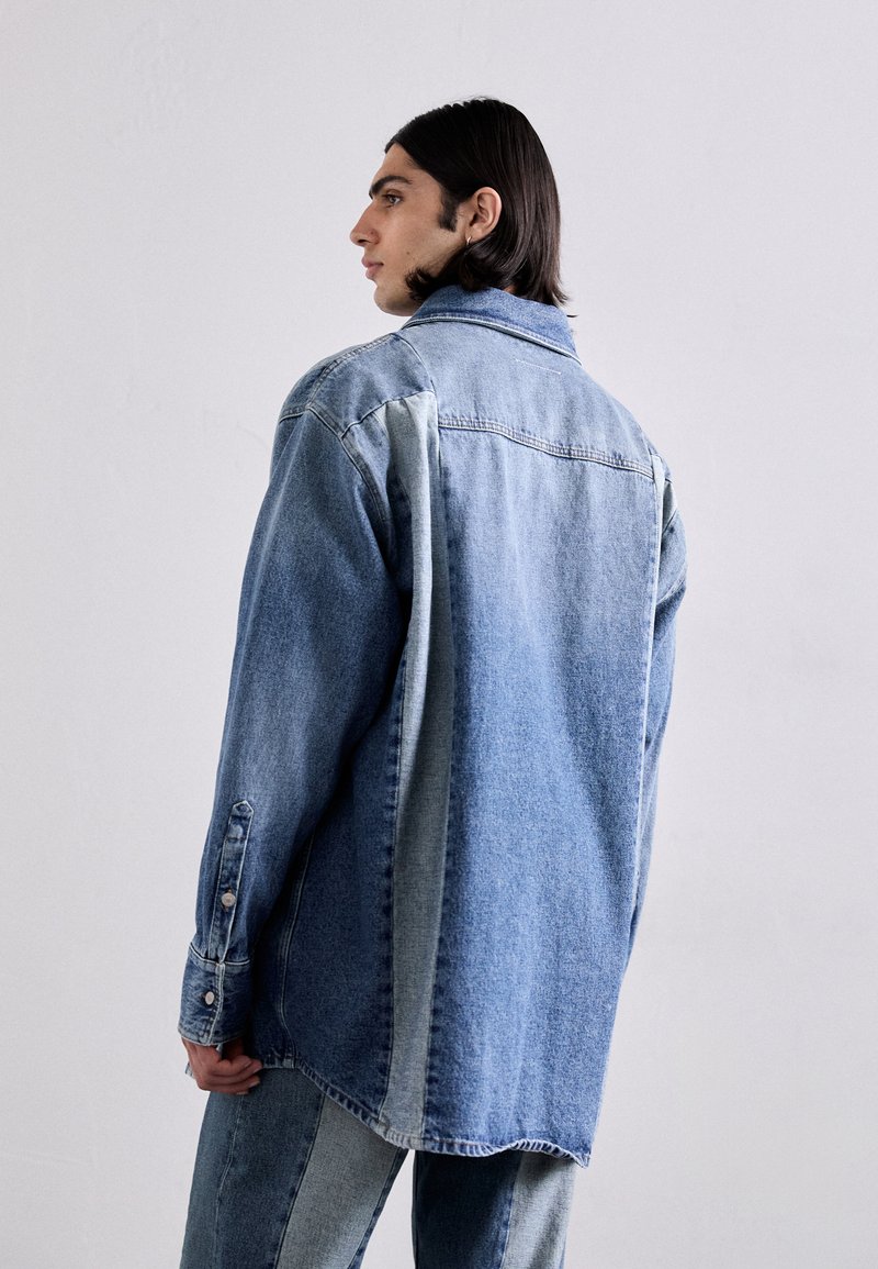 Camicia in denim azzurro chiaro con un pannello blu scuro a contrasto su un lato. Presenta maniche lunghe, polsini con bottone e una vestibilità comoda.