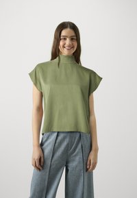 Grünes Oberteil mit hohem Kragen und kurzen Ärmeln, aus glattem Stoff, kombiniert mit hellblauen, gefütterten Hosen, beide vor einem neutralen Hintergrund präsentiert.