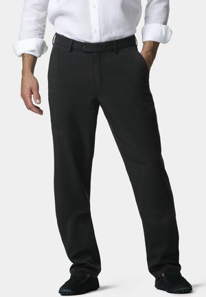 Homme portant un pantalon noir droit, une chemise blanche boutonnée aux manches retroussées, et des chaussures décontractées noires, debout avec une main dans la poche.