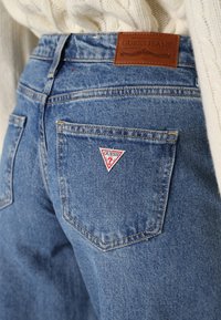 Vista trasera de unos jeans de mezclilla azul con parche de logo de Guess en el bolsillo trasero y etiqueta de cuero de la marca en la cintura, combinados con un suéter crema.