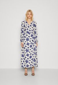 Robe à manches longues, décolleté en V, crème avec un imprimé floral bleu foncé. Tissu léger, corsage ajusté et jupe fluide. Accentuée par un détail à nouer.