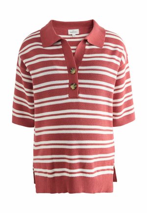 Femme aux cheveux noirs coiffés en chignon, portant un haut en maille à manches courtes rayé rouge et blanc ainsi qu'un pantalon assorti, debout de profil sur un fond uni.