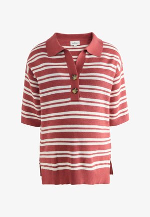 Moteris su tamsiais surištais plaukais, vilkinti raudonos ir baltos dryžuotos trumpomis rankovėmis megztu viršumi ir derančiomis kelnėmis, stovinti šonu priešais paprastą foną.