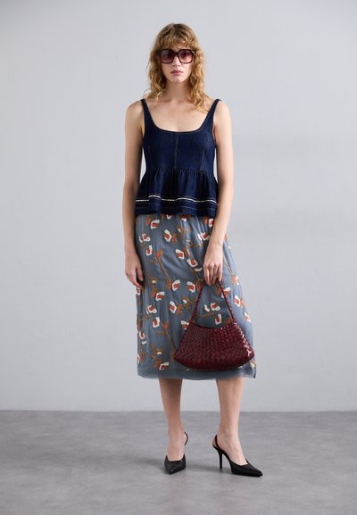 Denim-Peplum-Top mit quadratischem Ausschnitt, kombiniert mit einem blumengemusterten grauen Rock. Das Model hält eine gewebte burgunderfarbene Tasche und trägt schwarze Absatzschuhe.