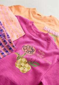 Három póló narancssárga és rózsaszín árnyalatokban, virágmintás grafikával, játékos részletekkel és különböző mintázatokkal, rétegezett elrendezésben.