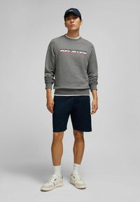 Grijze sweatshirt met meerkleurige letters, marineblauwe shorts en witte sneakers. Pet met logo in bijpassend grijs. Simpele, casual sportkleding.