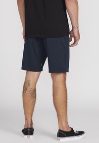 Granatowe szorty sportowe z lekkiego materiału, o luźnym kroju, z bocznymi kieszeniami, zestawione z czarnymi sneakersami i widocznymi tatuażami na nogach.
