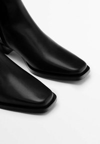 Stivali ankle in pelle nera con punta affilata, caratterizzati da una texture liscia e un tacco impilato, messi in risalto da una leggera lucentezza.