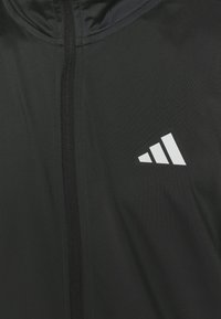 Felpa nera con zip realizzata in tessuto liscio, caratterizzata da un logo Adidas bianco con tre strisce. La zip scende verticalmente sul davanti.