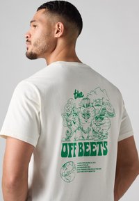 Homme portant un T-shirt blanc avec des légumes en cartoon verts et le texte "The Off Beets" dans le dos, regardant vers la gauche contre un fond gris.