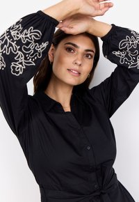 Vestido camisero negro con mangas largas y fruncidas. Presenta diseños florales bordados en hilo claro en las mangas. Tejido suave y ligero.