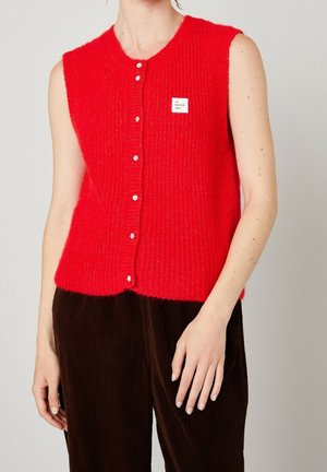 Gilet lavorato a maglia rosso senza maniche con bottoni bianchi e piccola etichetta bianca, indossato sopra pantaloni in velluto a coste marroni su una persona.